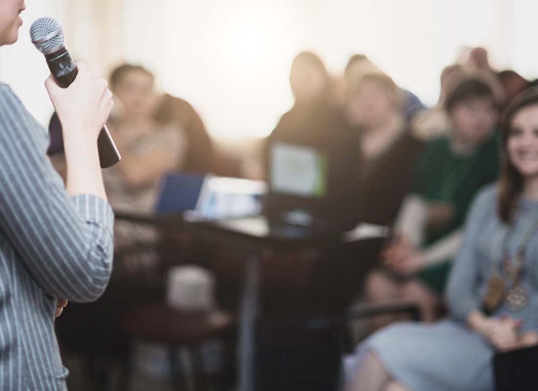Event Insurance - Focus On the Conference Speaker Using a Microphone and Standing in Front of a Group of People with the Audience Blurred in the Background