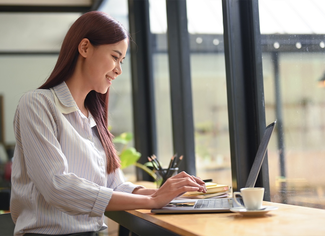 Service Center - Side View of a Cheerful Woman Sitting in a Modern Coffee Shop and Working Online With a Laptop